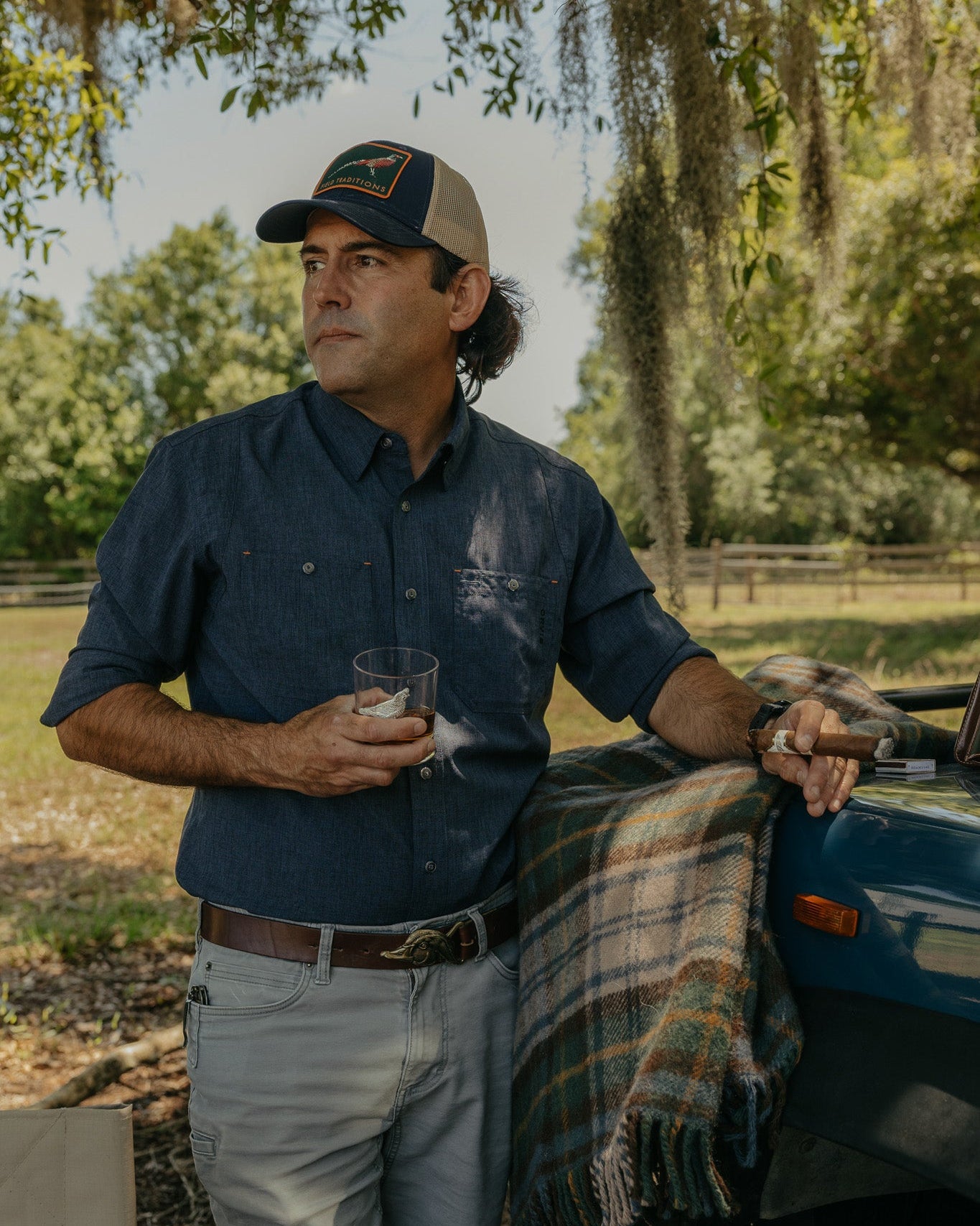 Man standing outdoors holding a glass, wearing a cap and blue shirt, with a plaid blanket on a vehicle.